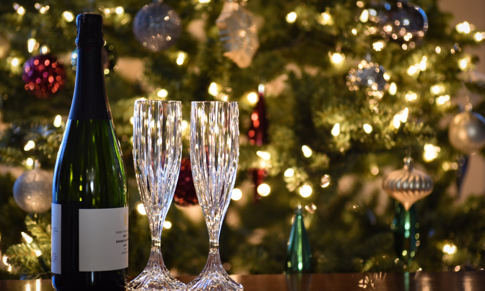 An image of a champagne bottle and two empty champagne glasses in front of a blurred out, buy brightly lit Christmas tree.