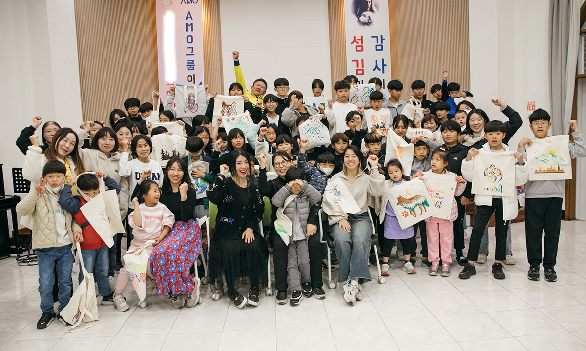 KKOOM volunteers and the children’s home staff and kids hold up their finished tote bags from an art event and smile at the camera.
