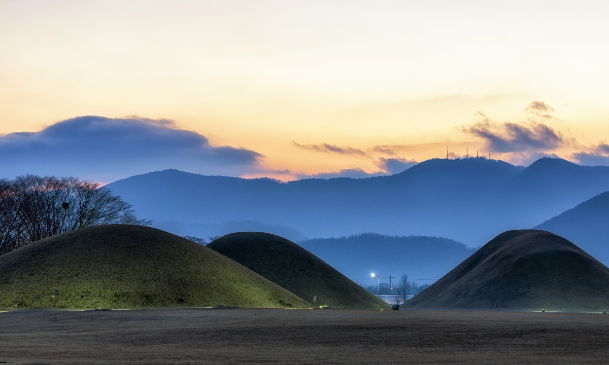 Alt Text: Large dark green burial mounds, with navy blue mountains the rising or setting sun on the horizon.