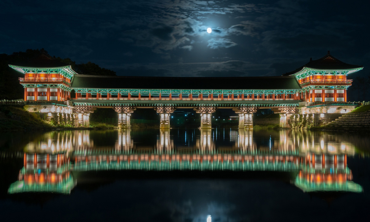 A full wide angle view of the Woljeonggyo Bridge lit up at night. The moon is bright and breaks through the cloudy night sky.