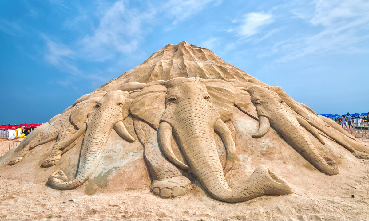 A large pyramid like sand sculpture with the frontal view of elephants charging forward on the base of the mound.