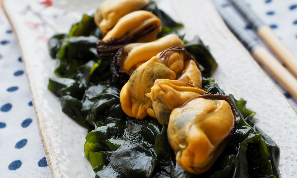 A row of raw muscles laid out on dark wet seaweed on a rectangle dish with chopsticks in the background.