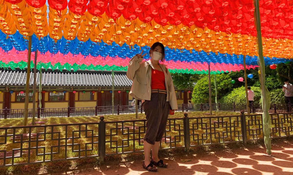 A woman stands underneath hundreds of colorful haanging lanterns that cast circle shadows on the ground.
