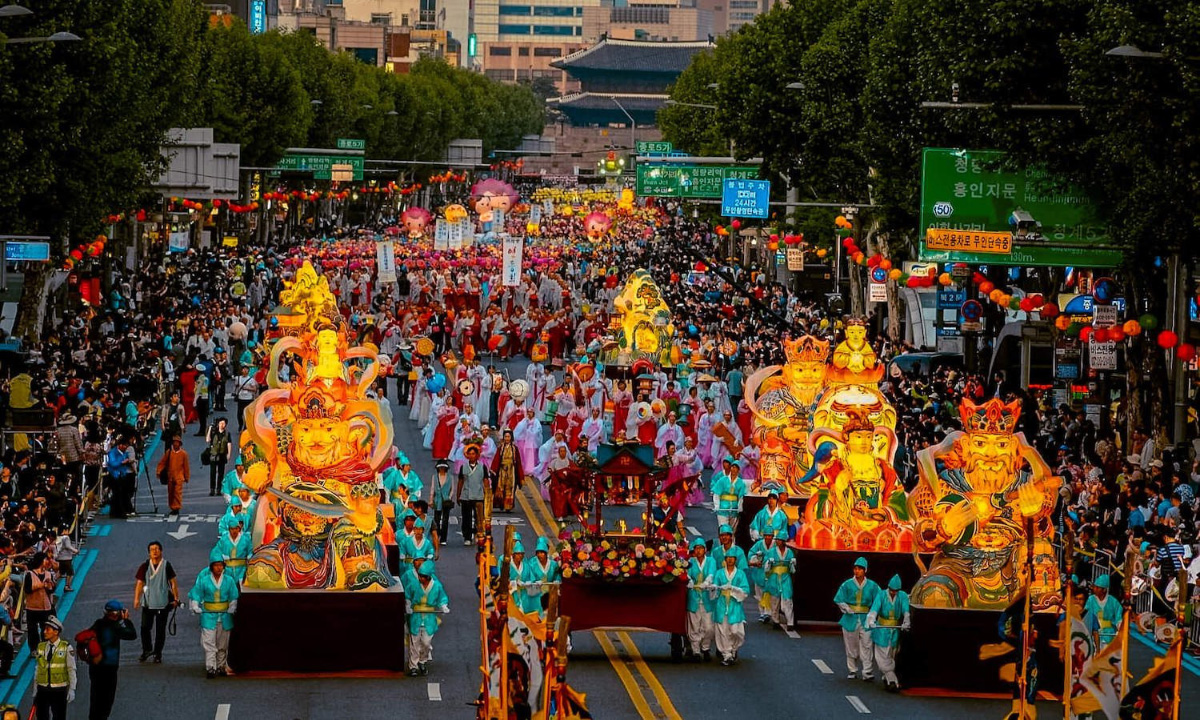 A large frontal view of an approaching parade of lanterns and lantern floats at dusk accompanied by people dressed in traditional Korean clothing. A large crowd of people watches the parade on either side.