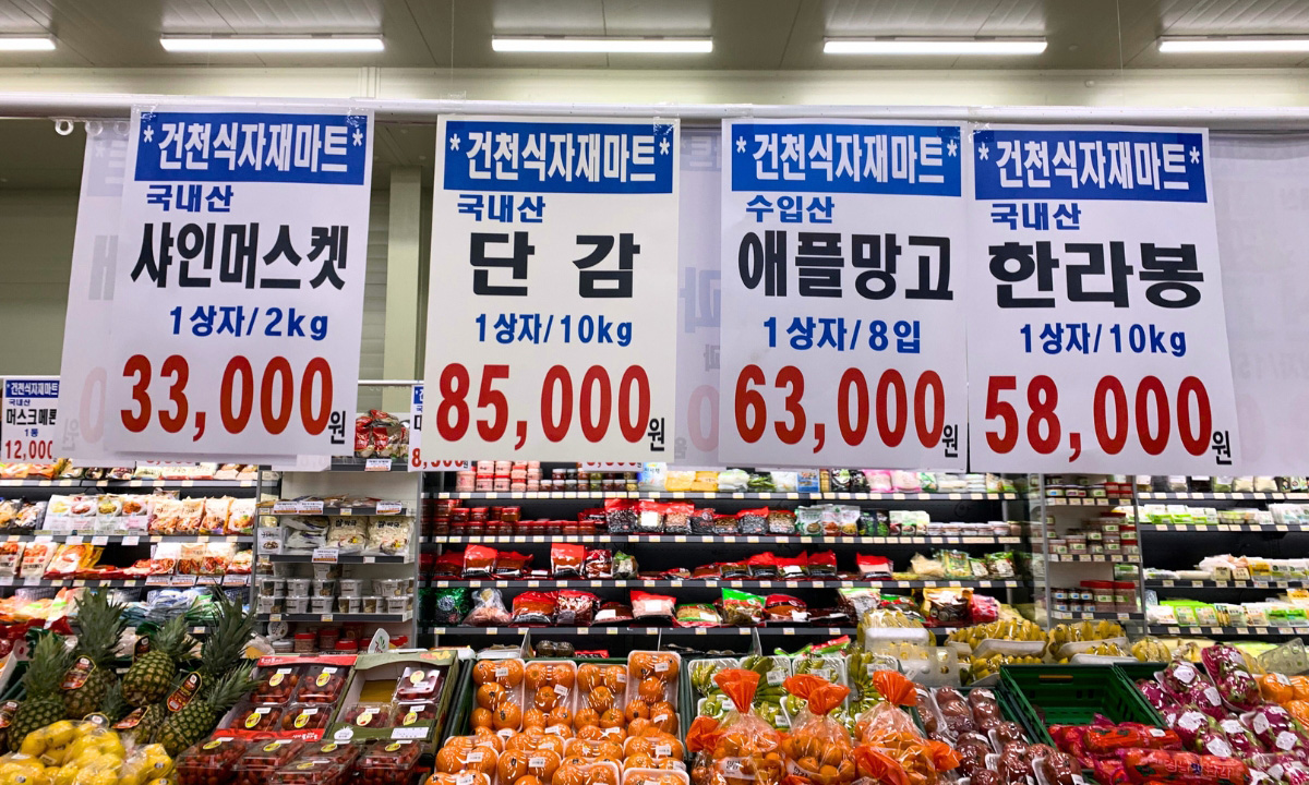 Inside look into produce stands of a grocery market with hanging signs in Korean that show how much the price is for the fruit below.