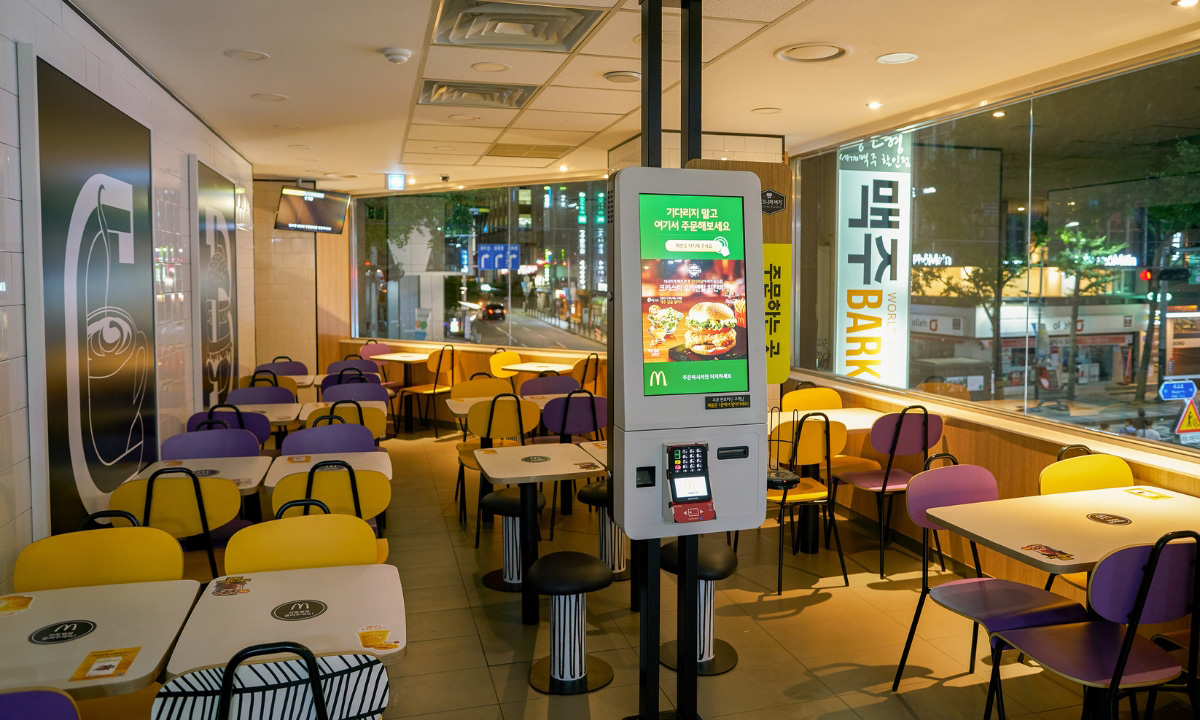The empty interior of a second floor McDonald’s with a kiosk in front of the tables and chairs.