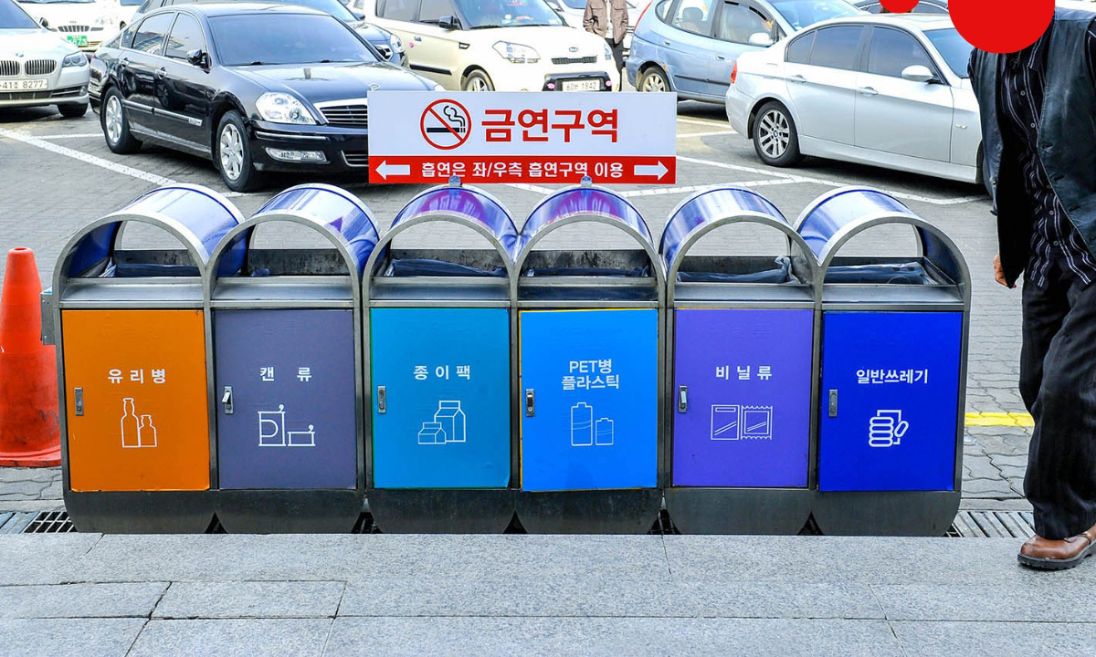 Six trash and recycling bins are near the curb of a parking lot with various labels in Korean.