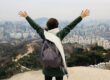 Woman faces her back to the camera and lifts up her hands on a mountain overlooking the city of Seoul.