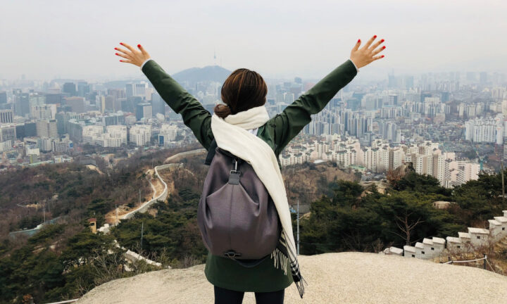 bok_travel-tips_travel-korea Woman faces her back to the camera and lifts up her hands on a mountain overlooking the city of Seoul.