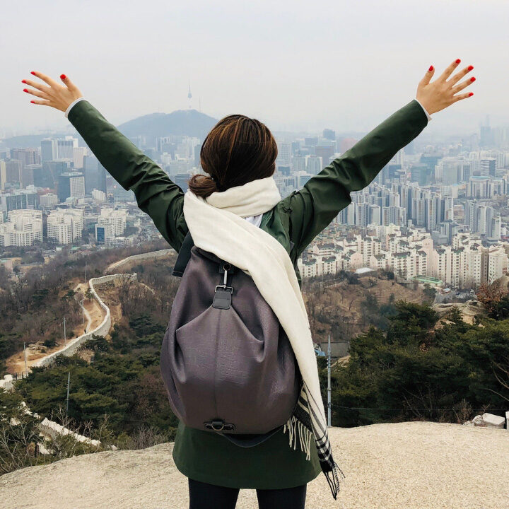 Woman faces her back to the camera and lifts up her hands on a mountain overlooking the city of Seoul.