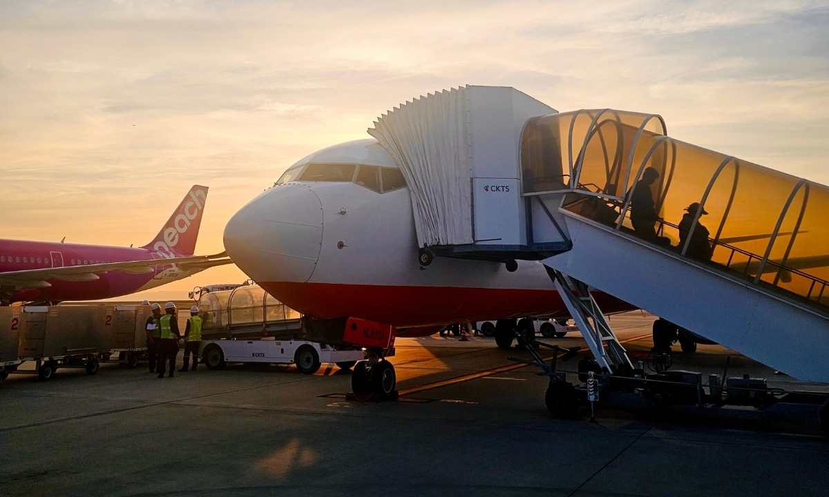 An airplane is parked on the tarmac at sunset, with passengers boarding via an exterior stairway. Ground crew members and another plane are visible in the background.