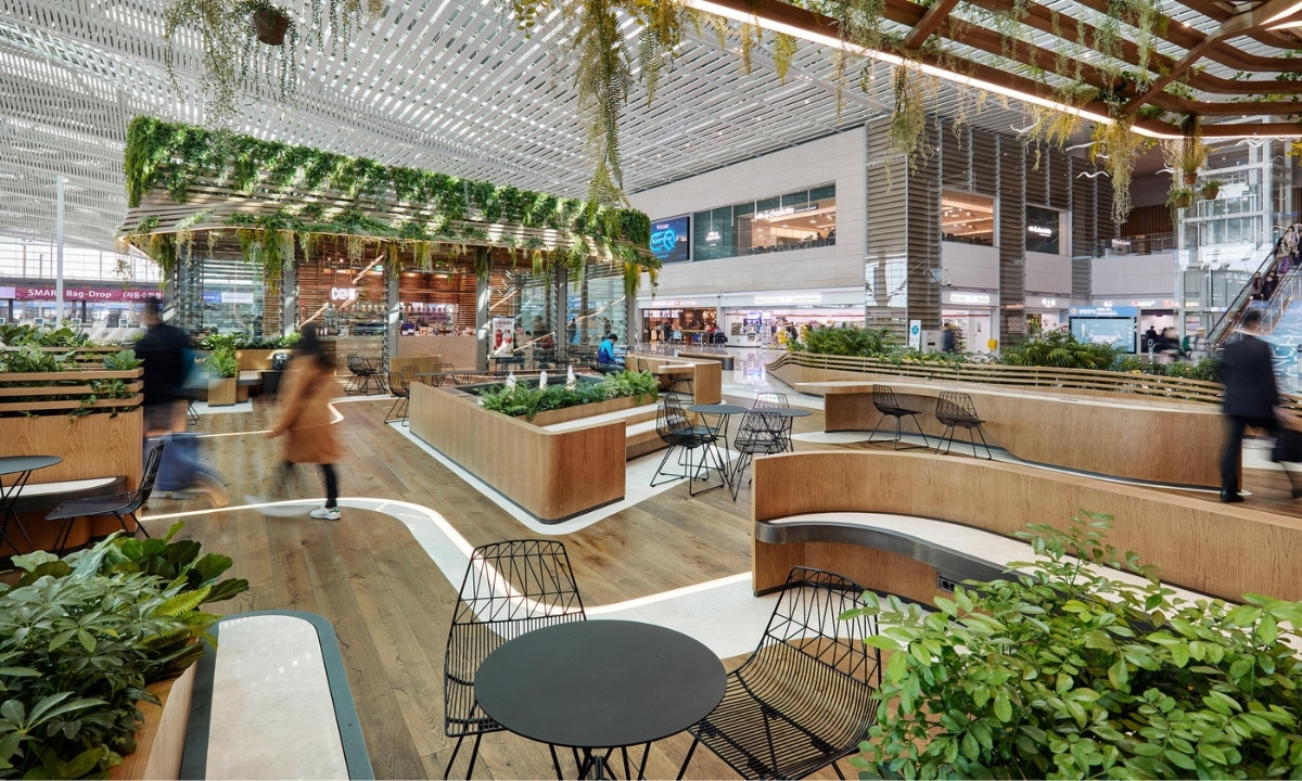 A spacious, modern indoor public area with seating and lush greenery, featuring hanging plants and a blend of wooden and metal elements, with people walking by.