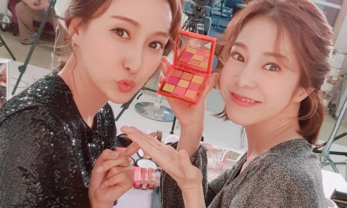 A Korean woman poses with Si-young for the camera showing off their makeup look and pink and gold eyeshadow palette. In the background is a makeup studio in Seoul.