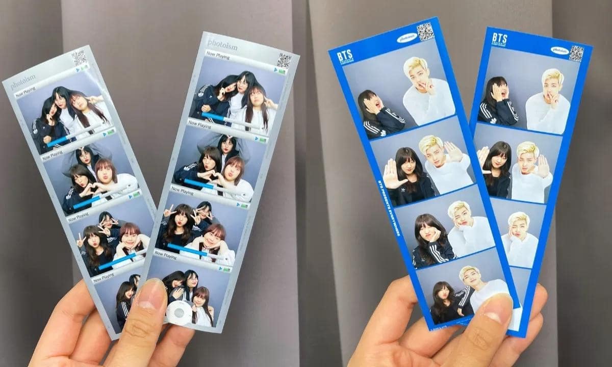 A person holds up two sets of Photoism Box x BTS photobooth 4-cut strips in-front of a gray curtain wall.