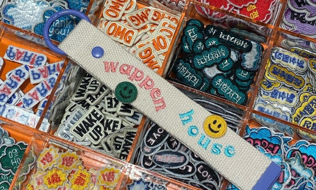 A white and purple Wappen House luggage tag is placed on top of rows and rows of press-on patches sorted into square plastic containers.