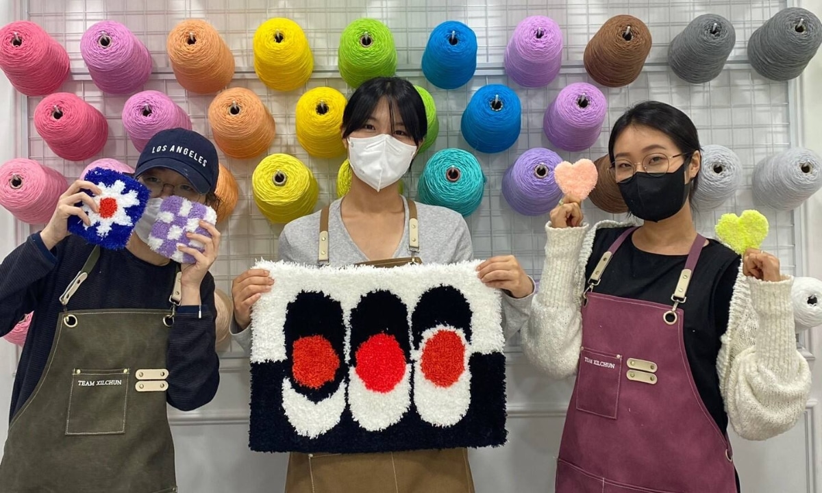 Three people in aprons hold up small rugs and tufting designs in-front of a walk of rainbow tufting string.