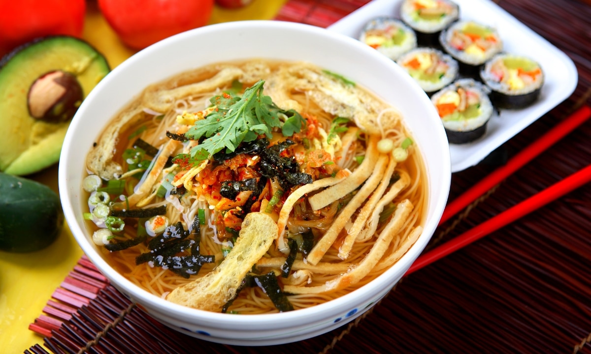 A large white bowl of janchi gulksu and six kimbap served on the side on a dark brown table.