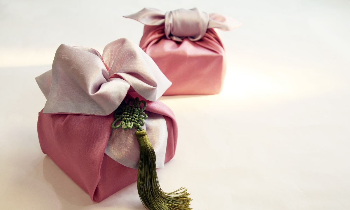Two pink bojagi wrapped square boxes with a green norigae tassle sitting on a white surface.