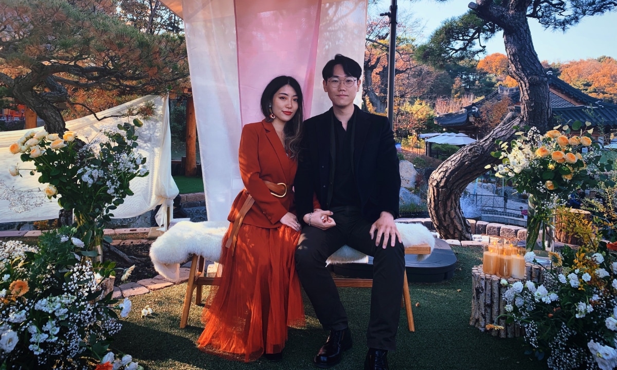 A couple sits in front of a beautiful Korean wedding backdrop in the midst of fall folliage and flowers.