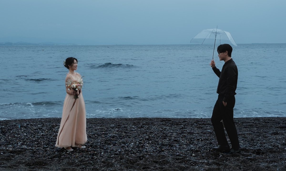 Couple standing across from one another on an overcast beach, the bride is wearing a light apricot colored dress and the groom is in all back and holding an umbrella.