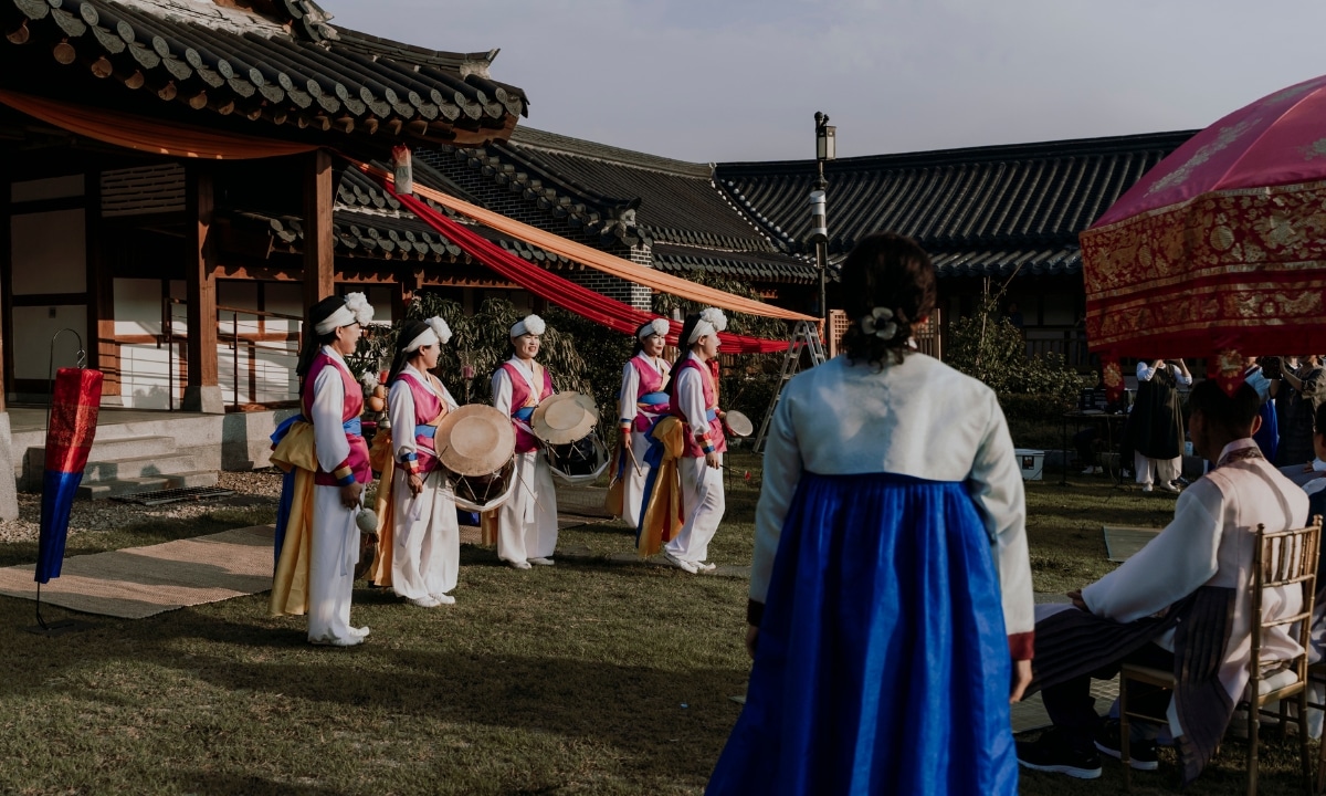 A bride and groom are seated on the far while a traditional Korean drum performance is peforming in front of them. They appear to be in a grassy hanok courtyard.