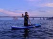 Swimmer wearing modest swimwear paddle boards in the sea under a sunset bright sky.