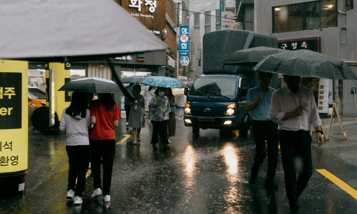People holding umbrellas walk through a busy street during a sudden downpour, typical of summer monsoon rains in Korean cities.
