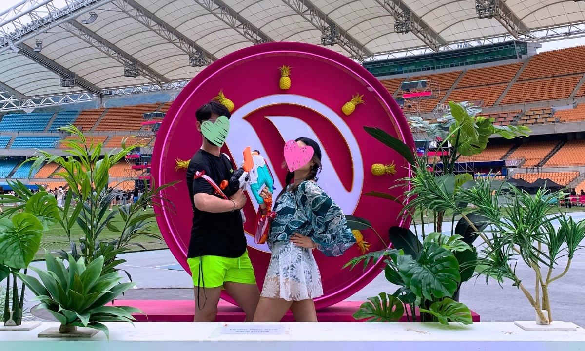 Two people posing for at a photo zone in front of the Waterbomb stage with performers and cheering crowds getting soaked in the background.